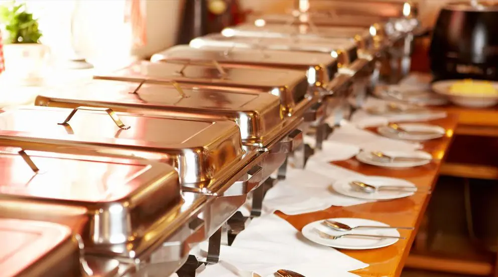 A row of shiny chafing dishes on a buffet table, set up with white plates and serving utensils in a warmly lit room.